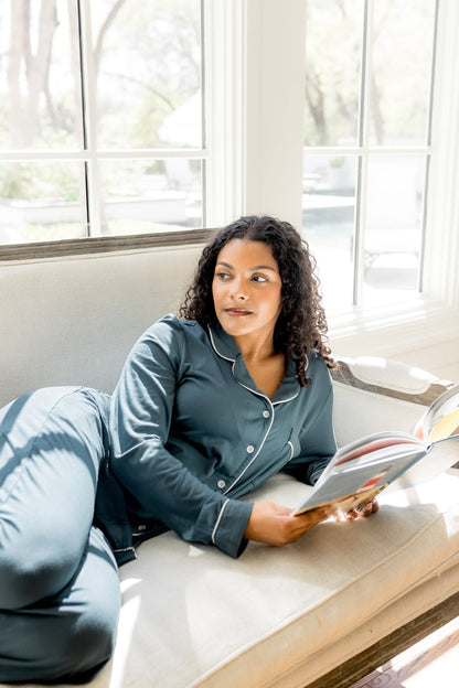 Female sitting on a couch underneath a window with a book in her hand wearing the Long-Sleeved Women's Pajama Set in Atlantic with Cloud Trim