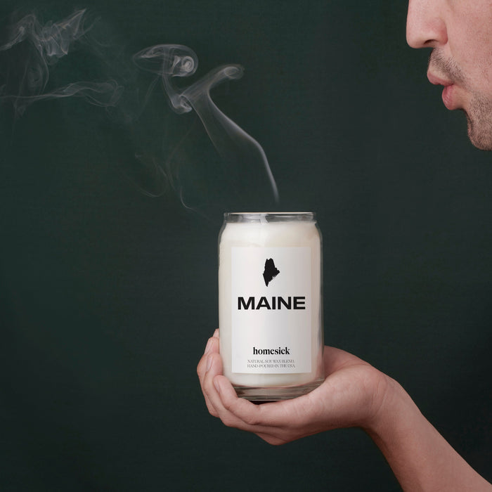 Someone holding a Maine homesick candle that they have just blown out so the smoke is coming up out of the candle.