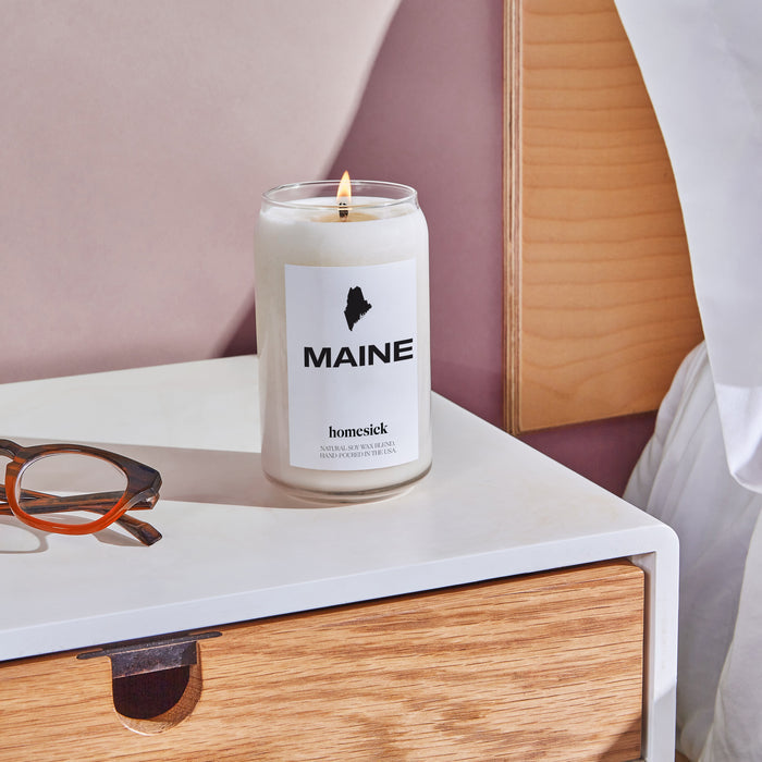 A bedside shot of the Maine Candle on a white side table with a wooden drawer.