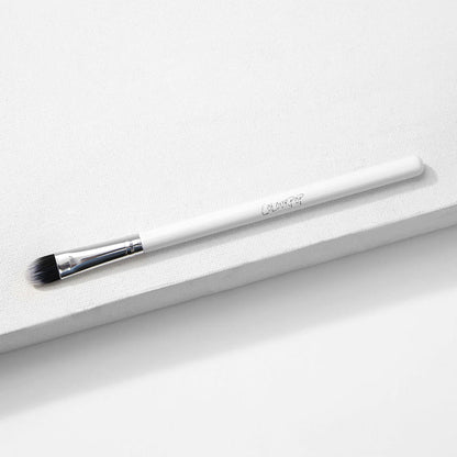 ColourPop Medium Shader Brush with flat oval, dual-toned black-to-white synthetic bristles and a silver ferrule on a white handle, resting on a white textured platform in studio lighting; makeup brush for precise eyeshadow packing and blending.