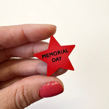 A hand with red nail polish holds a small red star-shaped magnet with the words 'MEMORIAL DAY' printed in black text against a plain off-white background.