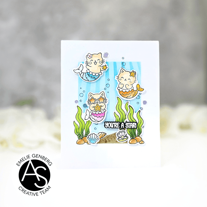 A white greeting card featuring a cute cartoon mermaid cat illustration with the text 'YOU'RE A STAR!' and underwater-themed details, shown against a soft blurred gray and white background. A circular logo in the lower left reads ALEX SYBERIA STAMPS DIES