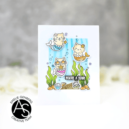 A white greeting card featuring a cute cartoon mermaid cat illustration with the text 'YOU'RE A STAR!' and underwater-themed details, shown against a soft blurred gray and white background. A circular logo in the lower left reads ALEX SYBERIA STAMPS DIES