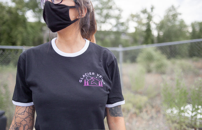 Black short sleeve shirt with white collar and white rimmed sleeves and GLACIER VEISN written in the top corner o the chest in white. There is a drawing of mountains below the text. An individual is modeling the shirt and standing in front of a fence with greenery and trees in the background.