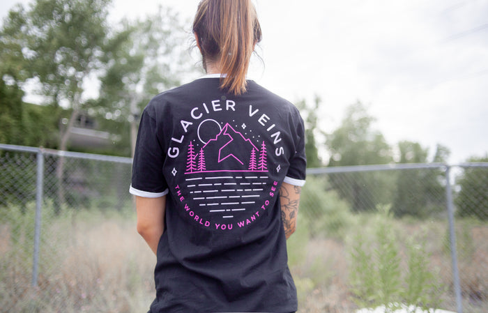Back of black short sleeve shirt with GLACIER VEINS written across the top. Below the text is a drawing of a mountain with a moon over it and stars in the sky. there are pine trees at the base of the mountain. Below the drawing is pink text that says THE WORLD YOU WANT TO SEE. An individual is modeling the shirt in front of a fence with greenery and trees in the background.