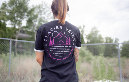 Back of black short sleeve shirt with GLACIER VEINS written across the top. Below the text is a drawing of a mountain with a moon over it and stars in the sky. there are pine trees at the base of the mountain. Below the drawing is pink text that says THE WORLD YOU WANT TO SEE. An individual is modeling the shirt in front of a fence with greenery and trees in the background.