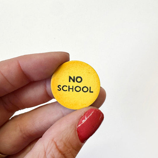 A hand holds a small round yellow magnet that reads 'NO SCHOOL' in black text against a plain white background.