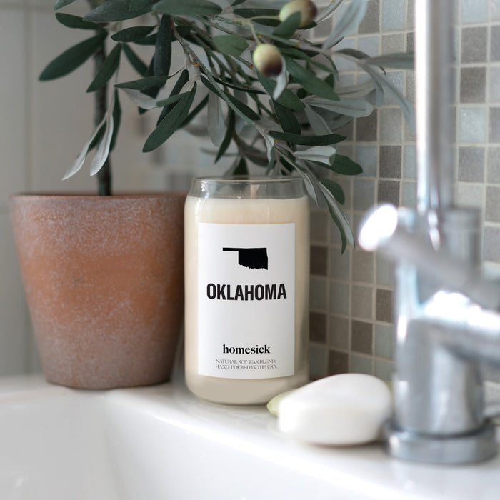 A close up shot of the Oklahoma candle on the edge of a white bathroom sink. There is a cermaic planter with an olive plat next to it.