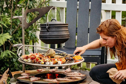 A woman cooking over a Breeo fire pit using a SearPlate Griddle, Outpost Grill, Kettle Hook, and Kettle