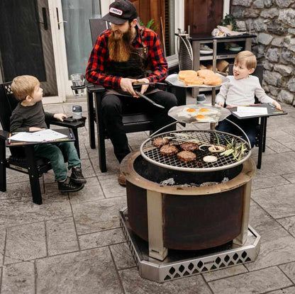 In this image a Dad is sitting around Breeo Firepit with his sons while grilling using an Outpost Grill and Outpost Sear Discs.