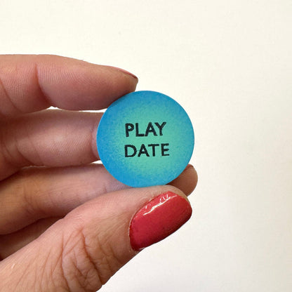 A hand holds a round blue magnet that reads 'PLAY DATE' in black text against a plain white background.