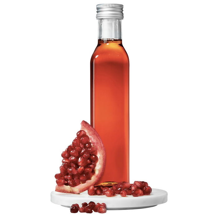 A clear bottle containing red pomegranate vinegar, with a silver cap, alongside a pomegranate fruit piece and scattered seeds.