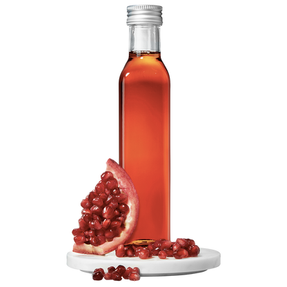 A clear bottle containing red pomegranate vinegar, with a silver cap, alongside a pomegranate fruit piece and scattered seeds.