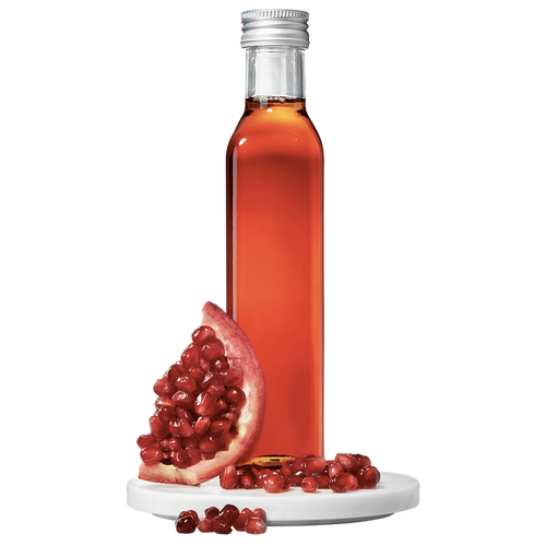 A clear bottle containing red pomegranate vinegar, with a silver cap, alongside a pomegranate fruit piece and scattered seeds.