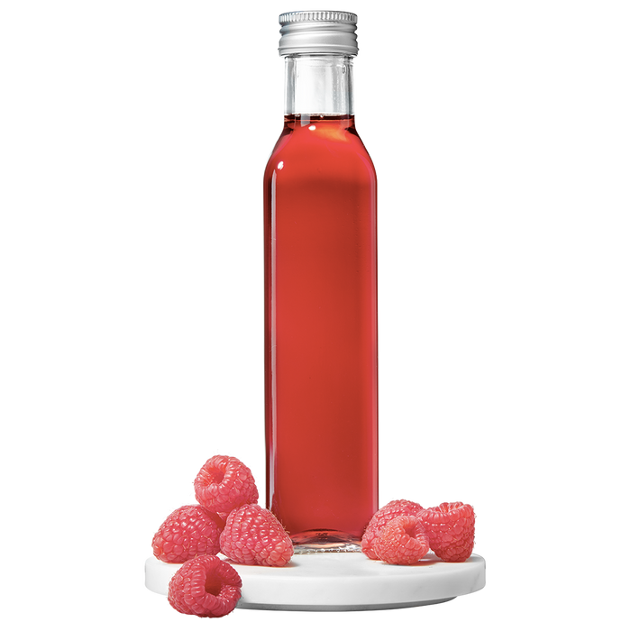A clear glass bottle filled with red raspberry vinegar, accompanied by fresh raspberries placed to the side.