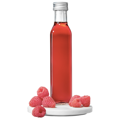 A clear glass bottle filled with red raspberry vinegar, accompanied by fresh raspberries placed to the side.