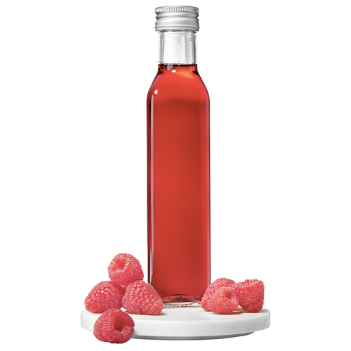 A clear glass bottle filled with red raspberry vinegar, accompanied by fresh raspberries placed to the side.