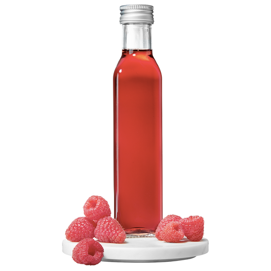 A clear glass bottle filled with red raspberry vinegar, accompanied by fresh raspberries placed to the side.