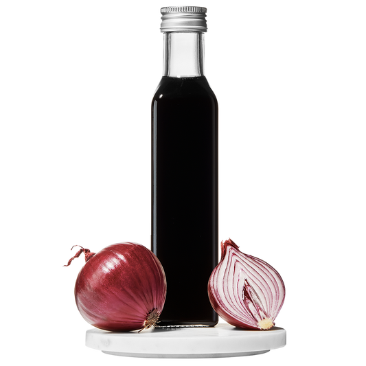 A clear glass bottle of Red Onion Balsamic Vinegar with a cap on top. A whole red onion and a slice of red onion are placed beside the bottle.