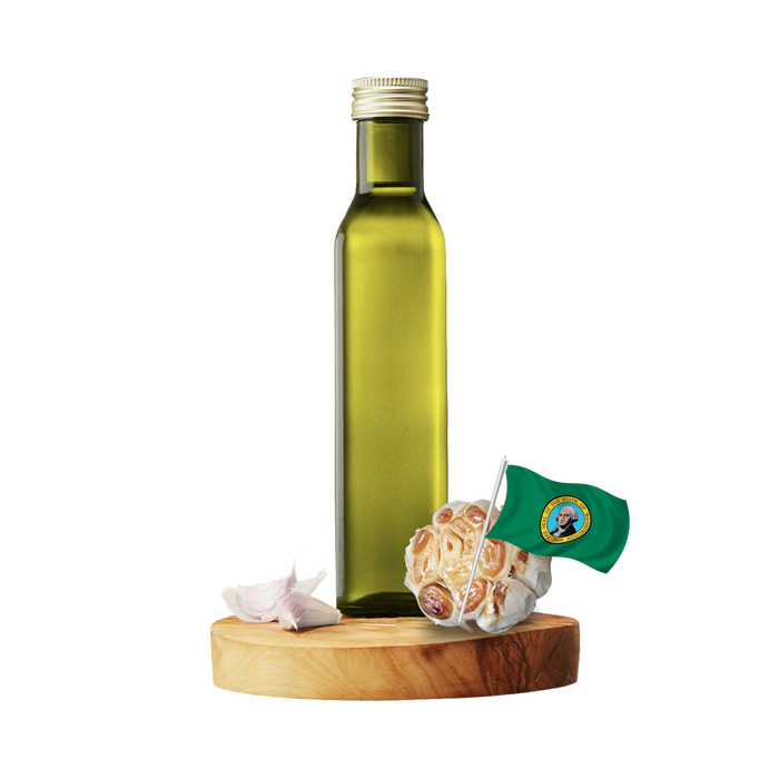 A tall olive oil bottle with a gold cap stands on a round wooden board, alongside garlic bulbs and a small green flag with a seal, all against a plain white background.