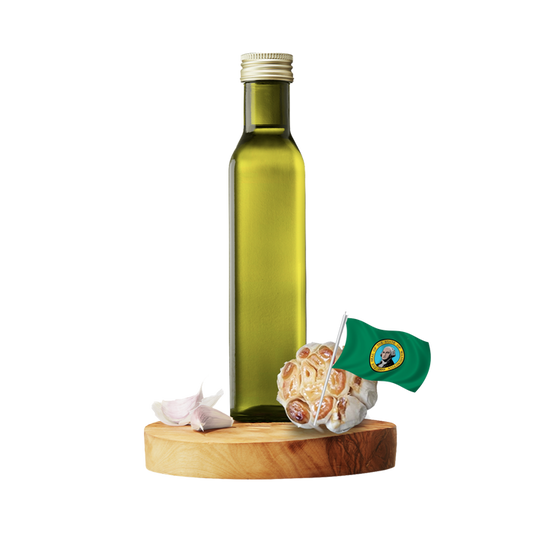 A tall olive oil bottle with a gold cap stands on a round wooden board, alongside garlic bulbs and a small green flag with a seal, all against a plain white background.