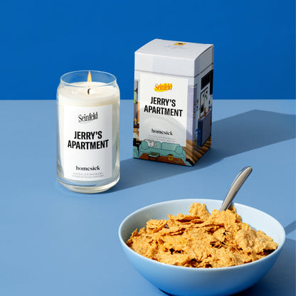 A stylized studio shot of the Jerry's Apartment candle from Homesicks Seinfield collection. There is a bowl of cereal in the lower right corner of the image. In the back left and middle is the Jerry's Apartment candle and packaging. They are shot on a light blue surface with a darker blue background.