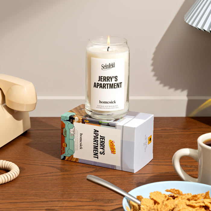 A stylized lifestyle shot of the Jerry's Apartment candle displayed on top of the packaging. Around the corners of the image are props of ones house.