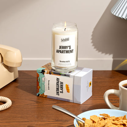 A stylized lifestyle shot of the Jerry's Apartment candle displayed on top of the packaging. Around the corners of the image are props of ones house.