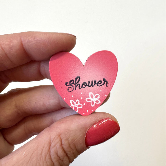 A hand  holds a small pink heart-shaped magnet that says 'Shower' in black script with white flower illustrations, against a plain off-white background.