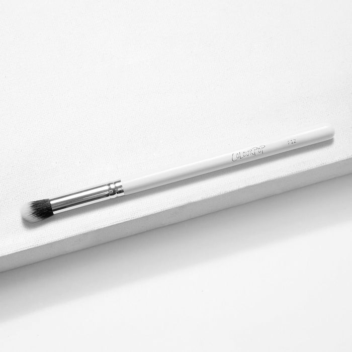ColourPop Small Detail Brush lying diagonally on a white textured backdrop, showcasing tapered black-to-white synthetic bristles, a silver ferrule, and a slim white handle; precision makeup brush for detailed eyeshadow and smudging.