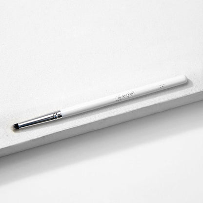 ColourPop Small Pencil Brush makeup brush lying diagonally on a white textured block, showing a slim white handle, silver ferrule, and small tapered bristles for precise eyeshadow application and smudging.
