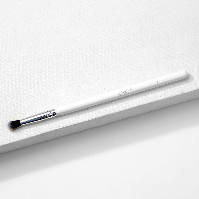 ColourPop Small Tapered Brush resting diagonally on a white textured platform, featuring a slim white handle, chrome ferrule, and small tapered black-and-white bristles for precise eyeshadow blending and detail work.