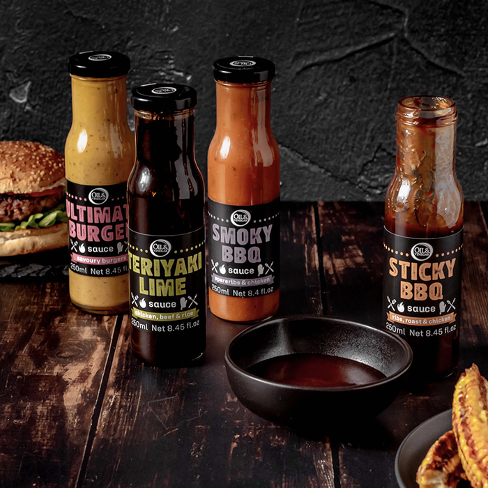 Fours bottles of sauces made for barbecuing Ultimate burger sauce, Teriyaki Lime Sauce, Sticky BBQ sauce, and Smoky BBQ sauce all on a wooden board with a burger in the background and small bowl of sauce in the foreground