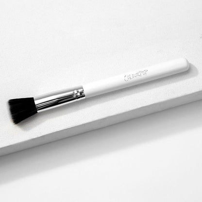 ColourPop stippling makeup brush resting diagonally on a white textured block, featuring duo-fiber black-and-white bristles, a shiny silver ferrule, and a white handle with the ColourPop logo.