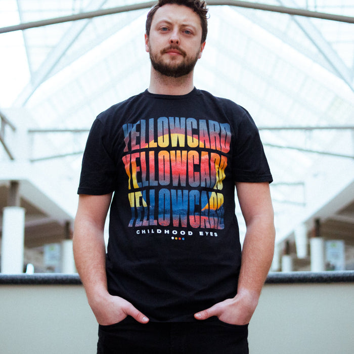 A bearded man stands indoors, wearing a Yellowcard Sunset T-Shirt with bold, colorful Yellowcard lettering over CHILDHOOD EYES. The vibrant Comfort Colors fabric contrasts with glass and metal architectural details.