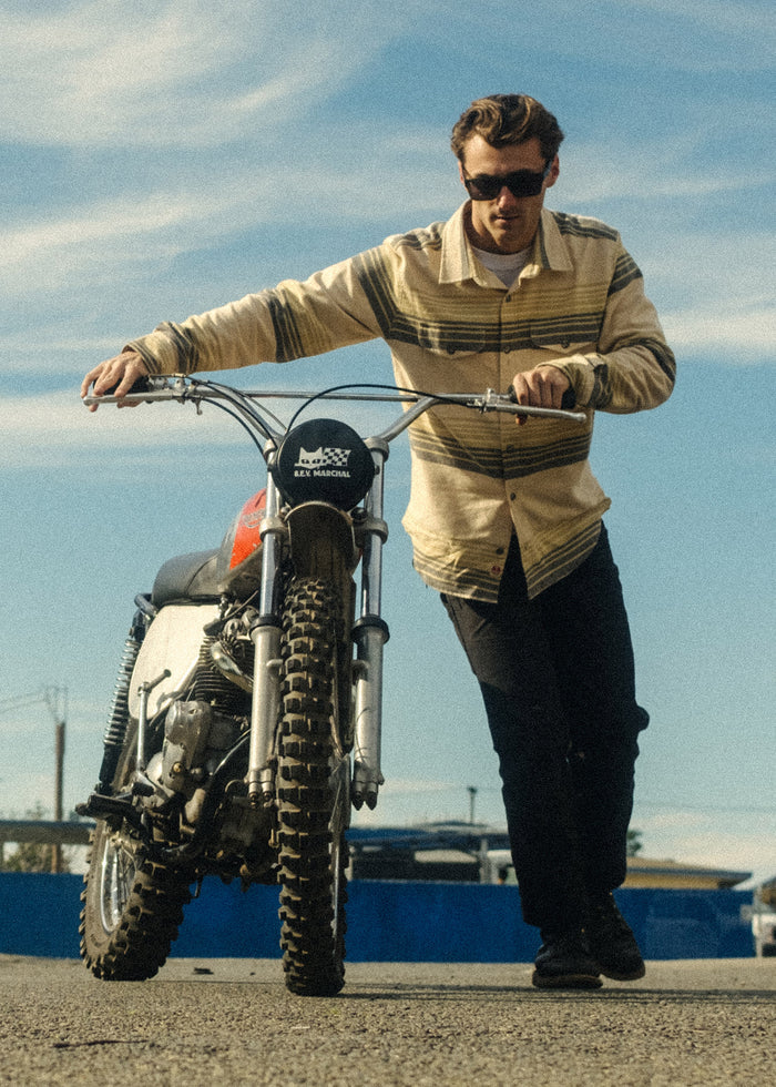 Man in sunglasses pushes a dirt bike on a sunny day, wearing a striped shirt. Blue sky and track barriers in the background.