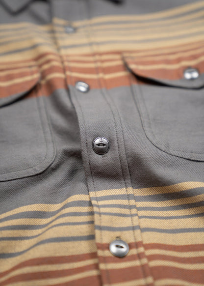 Close-up of a striped shirt with brown, yellow, and gray hues, featuring metal buttons and chest pockets for a casual look.