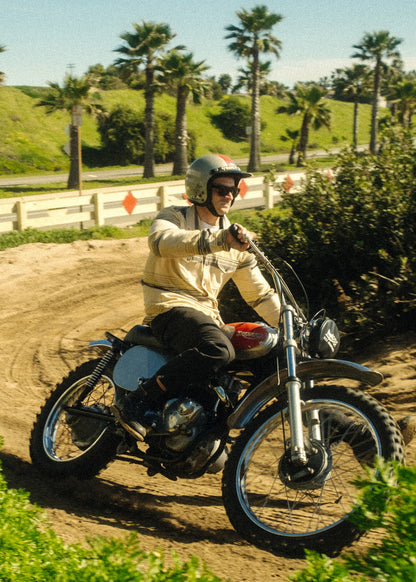 Man rides vintage motorcycle on a dirt path, surrounded by palm trees and greenery, under a clear blue sky.