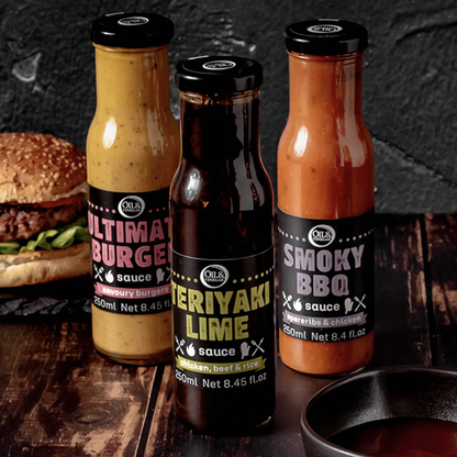 three jars of sauces made for barbecuing Ultimate burger sauce, Teriyaki Lime Sauce, and Smoky BBQ sauce all on a wooden board with a burger in the background