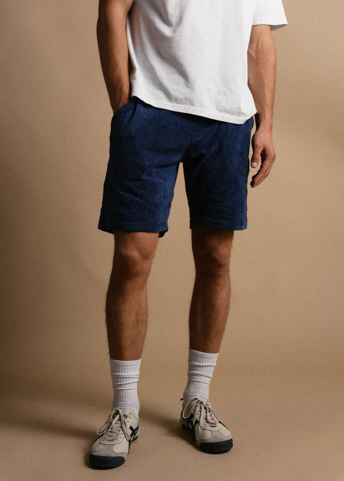 Man in white shirt, blue shorts, and sneakers stands on beige background, hands in pockets, showcasing casual summer style.