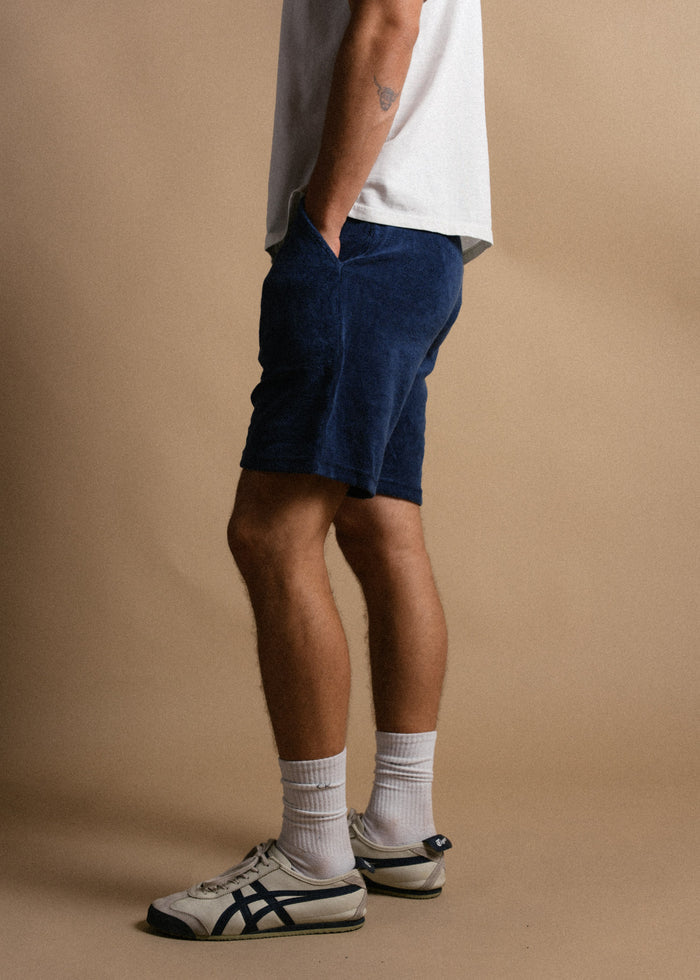 Man in white shirt, blue shorts, and sneakers stands against a beige background, hands in pockets, showcasing casual style.