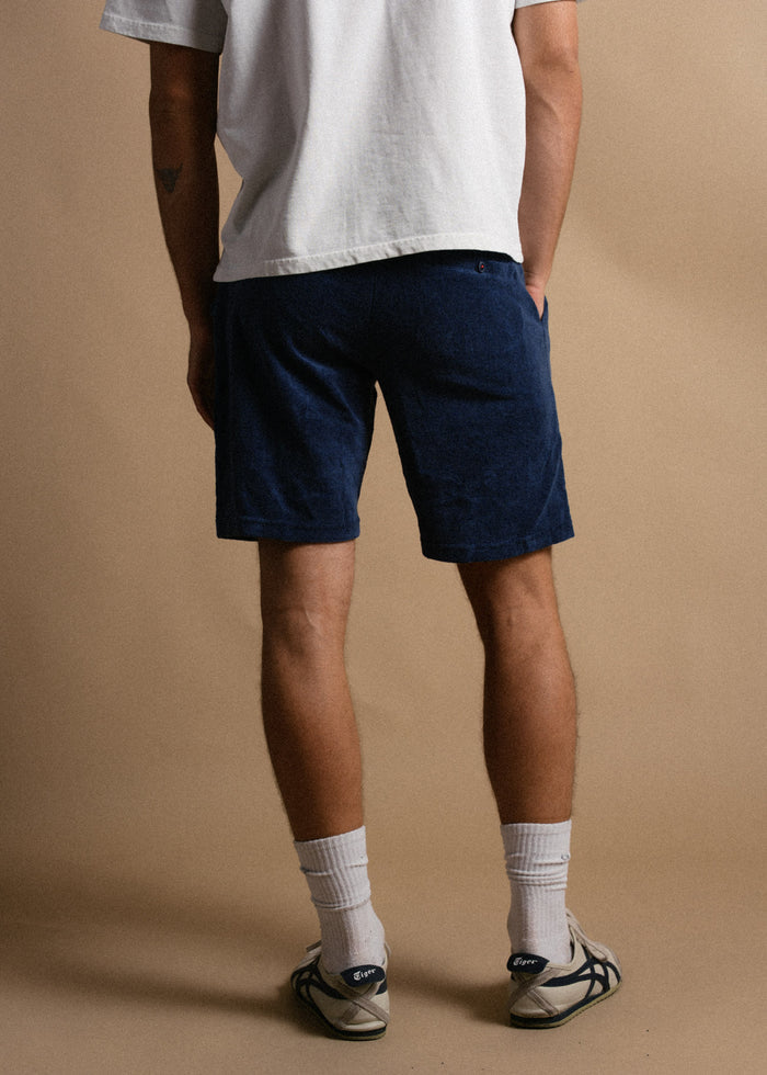 Man in white shirt and blue shorts stands against a beige background, wearing white socks and sneakers.