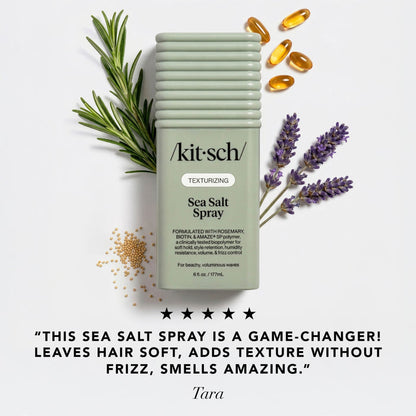 A bottle of kitsch Texturizing Sea Salt Spray centered on a light gray background, surrounded by rosemary sprigs, lavender stems, scattered sea salt grains, and amber capsules.