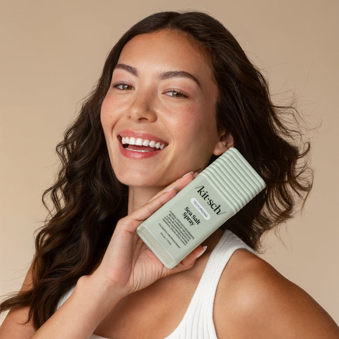 Smiling woman with voluminous, wavy dark hair holding a KITSCH Texturizing Sea Salt Spray bottle