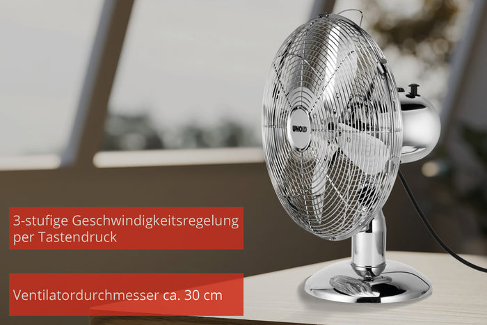 Der Unold TISCHVENTILATOR CHROME, ein 30 cm großer Chrom-Tischventilator, steht auf einer Holzoberfläche. Der deutsche Text zeigt die 3-stufige Steuerung und die Größe. Ein sanft verschwommener Hintergrund lässt Licht und Grün erkennen.