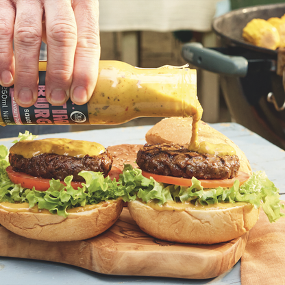 Burgers with lettuce, tomatoes, and buns on an olive wood board with a hand dressing them with the Oil & Vinegar branded Ultimate burger sauce