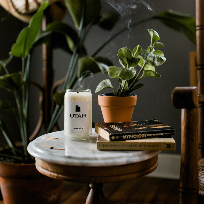 The Utah candle shot on a marble side table that has books and a potted plant on it as well. In the back is a blurred fig tree in a dark room.