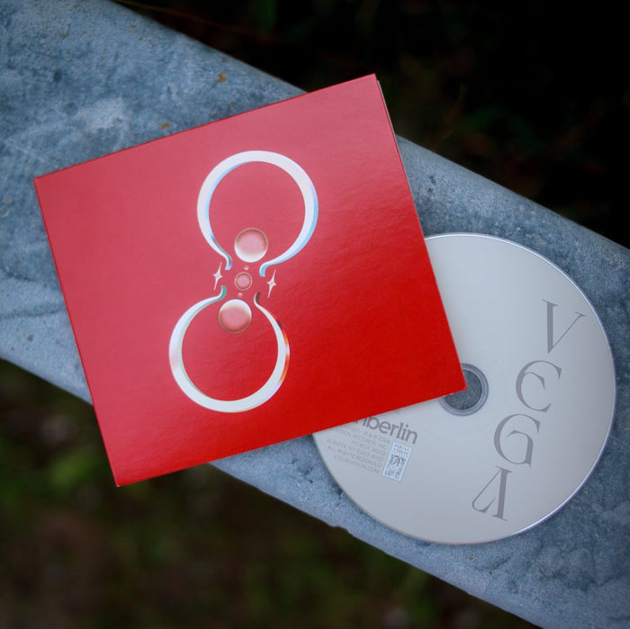 A red CD showcases two silver/gold rings facing each other with sparkles around them. A white CD that says "Vega" lies on top of a metal railing outside.