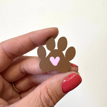 A hand with red painted fingernails holds a small brown paw print magnet with a pink heart in the center against a plain off-white background.