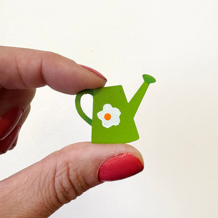 A hand holds a small green watering can-shaped magnet featuring a white flower with a yellow center, against a plain white background.